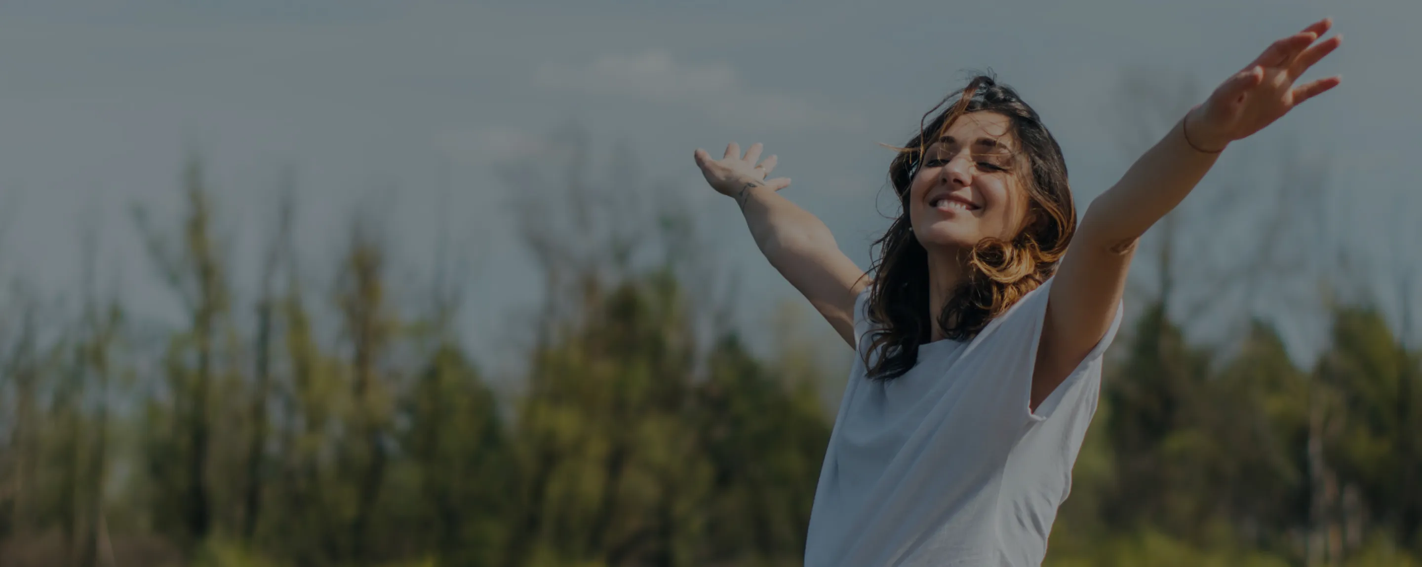 A photo of a woman with her arms spread wide and her eyes closed in nature.
